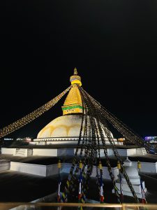 
Illuminated stupa at night with colorful prayer flags extending from its golden spire. The stupa has large painted eyes and a peaceful ambiance.