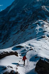 A lone hiker wearing a bright red backpack and holding a trekking pole walks along a narrow, snowy ridge path. Other hikers are visible in the distance on the snow-covered mountain slope, which is cast in deep blue shadows and bright sunlight.