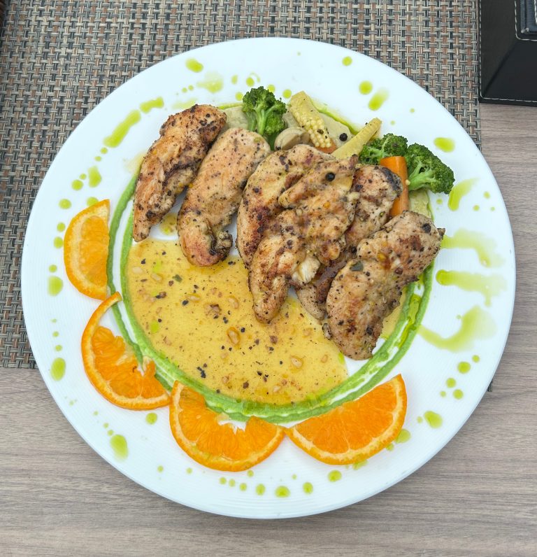 Grilled chicken pieces on a white plate with a yellow sauce, garnished with sliced oranges and vegetables, on a woven placemat.