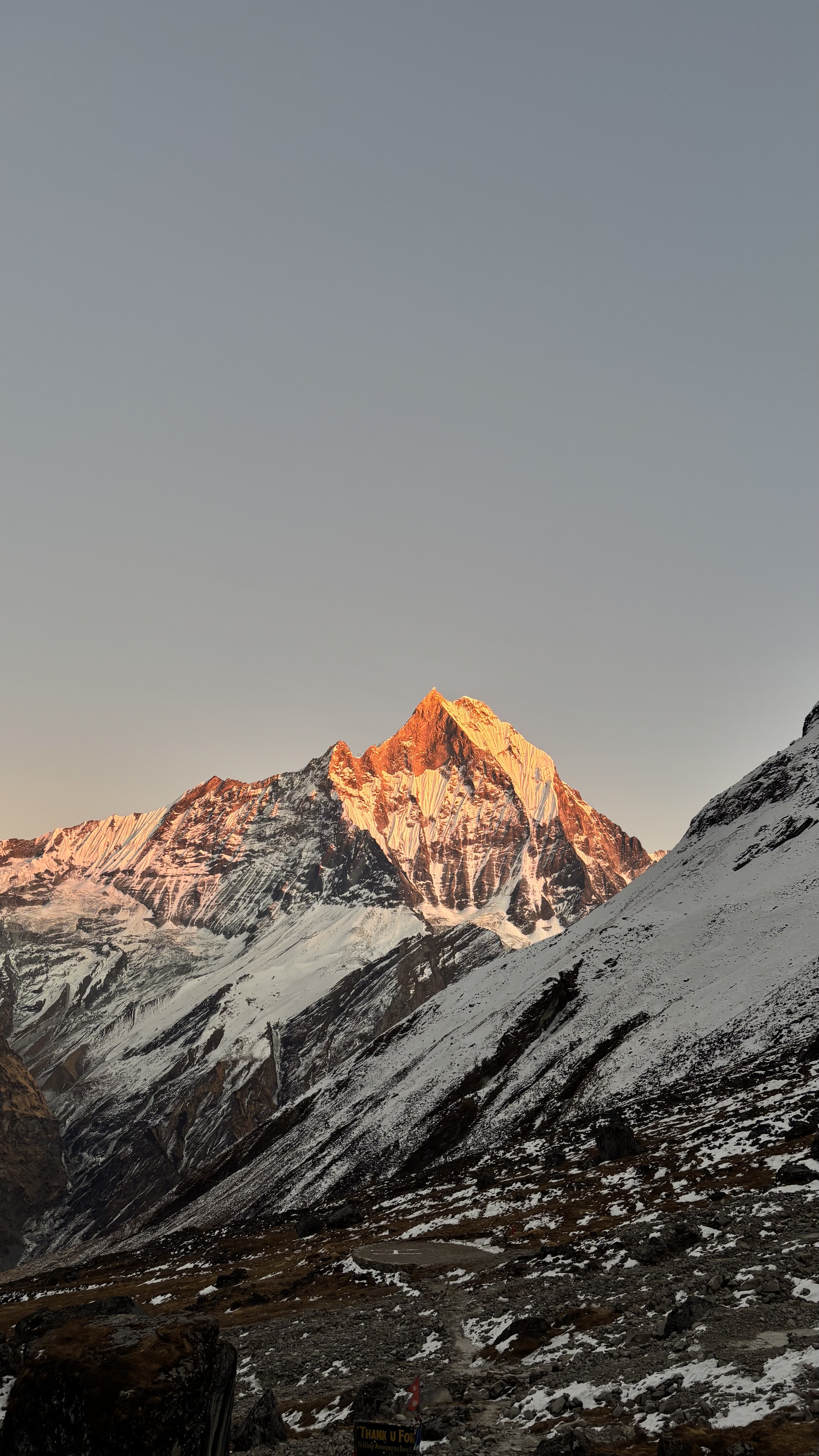 A snow-covered mountain glows in sunrise light beneath a soft pastel sky. Rocky ground and a partially visible welcome sign sit in the foreground of this quiet, high-altitude scene.
