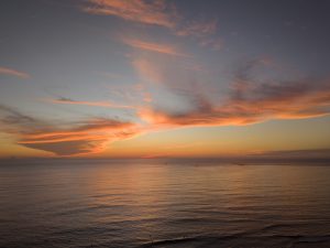 A wide ocean sunset with soft orange and pink clouds stretching across the sky. The calm water reflects the warm evening light, with small waves and tiny figures visible near the shoreline.