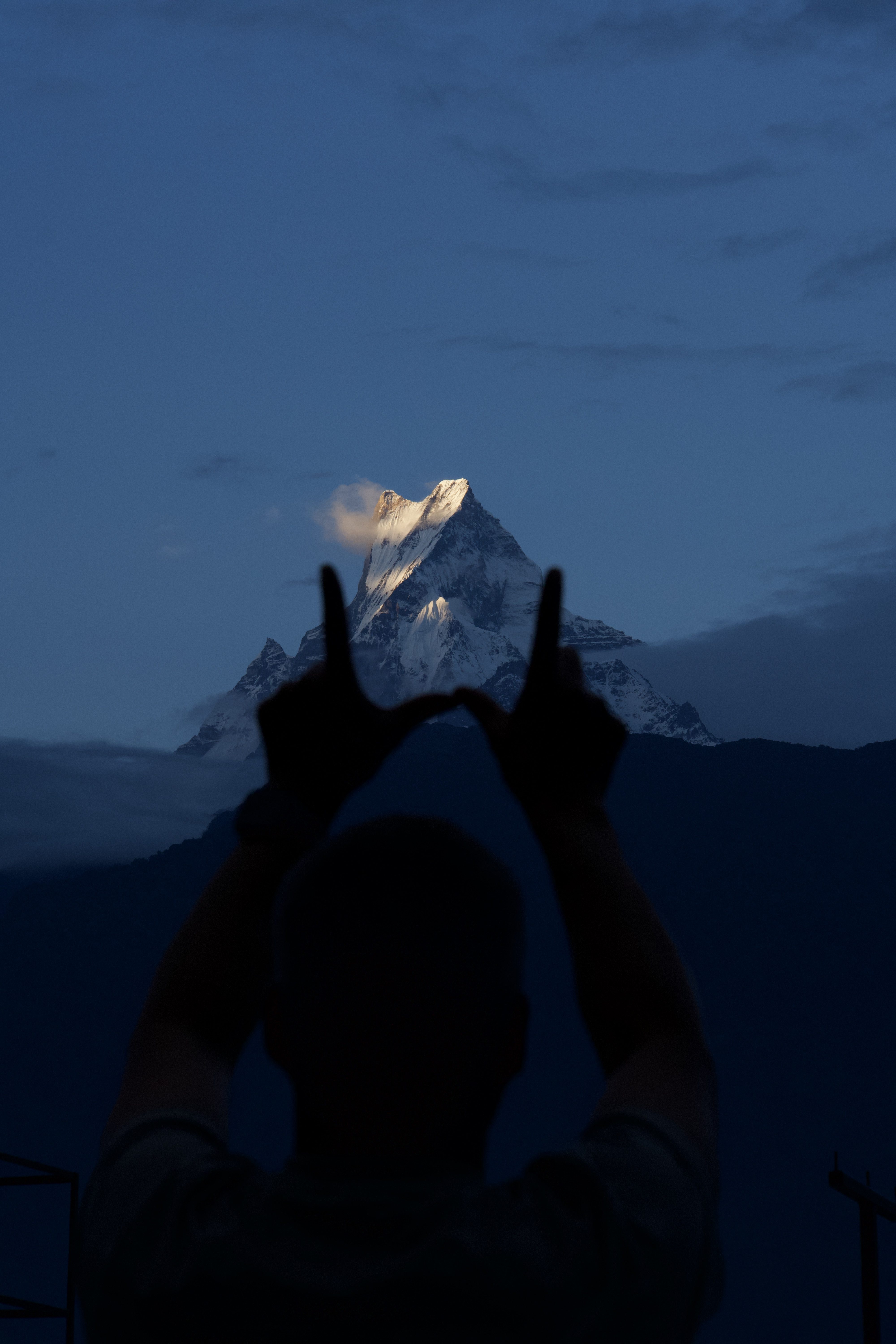 Silhouette of a person making a hand gesture with the majestic Fishtail Mountain in the background, viewed from Ghandruk, Nepal, during dusk under a calm blue sky.