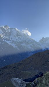 A trekker lying on a rocky ridge resting in front of a massive snow covered Himalayan peak under a clear blue sky. 