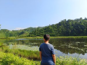 A young person stands contemplatively at the edge of a lily-covered lake, gazing toward snow-capped Himalayan peaks beyond green hills.