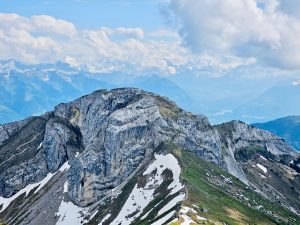 A dramatic alpine landscape showcases the rugged cliffs of Mount Pilatus, with patches of snow and distant mountain ranges stretching beneath a sky filled with clouds. The sweeping panorama highlights the grandeur of the Swiss Alps and the beauty of high-altitude scenery. Captured at Pilatus Kulm, Lucerne, Switzerland.