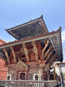 A richly decorated two-tiered Newari pagoda temple facade with elaborately painted wooden struts depicting deities, prayer wheels around the base, and colorful carved details, Kathmandu, Nepal. 
