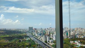 City skyline view of Mumbai from a high-rise window, showing skyscrapers, a busy highway, and green areas under a partly cloudy sky.