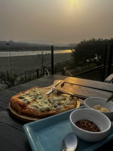 Pizza slices on a wooden board with spices and fork on a patio table. A serene riverside view is seen under a golden sunset sky in the background.
