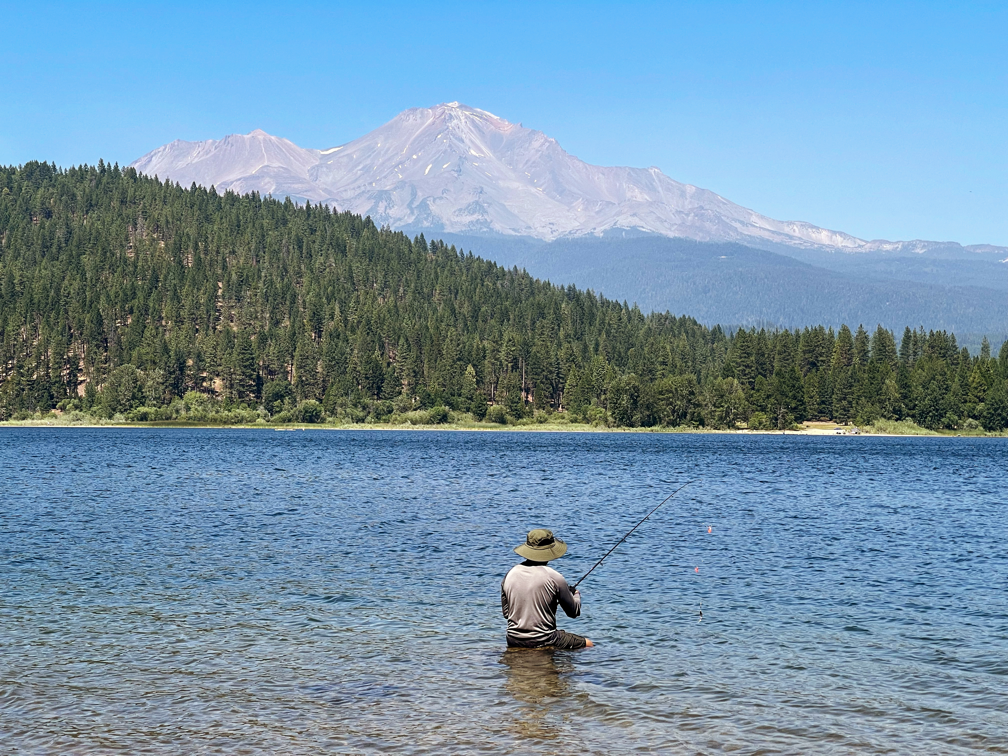 An angler sits fishing in a wide blue lake surrounded by a dense forest of evergreen trees. In the distant background, a massive, snow-dusted volcanic mountain peak rises into a clear sky.