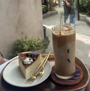 A glass of iced coffee on a coaster sits beside a slice of cheesecake topped with cream and seeds on a wooden tray. A person walks past outside.