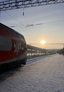 A red and gray train at a snowy station during sunrise, with overhead cables and an empty platform. 