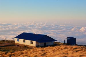 A lone house with a blue roof sits on a grassy hilltop above a sea of clouds at sunset, conveying tranquility and isolation in the mountainous landscape.
