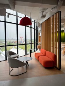 Modern lounge with a bright orange couch, gray chairs, a black coffee table, and large curved windows overlooking greenery.