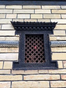 Intricately carved blackened wooden window with lattice grille and pagoda-style canopy on a traditional Newari stone brick temple wall, Nepal. 
