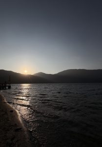 
Sunset over a lake with gentle waves, sand in the foreground, mountains in the distance, and a small dock with a flag on the left.