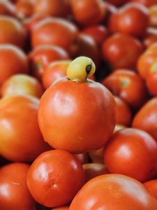A ripe red tomato stands out among a pile of fresh tomatoes, topped with a small yellow fruit that resembles a natural crown. The vibrant colors and shallow depth of field create an eye-catching composition. Captured at Perumanna, Kozhikode, Kerala.