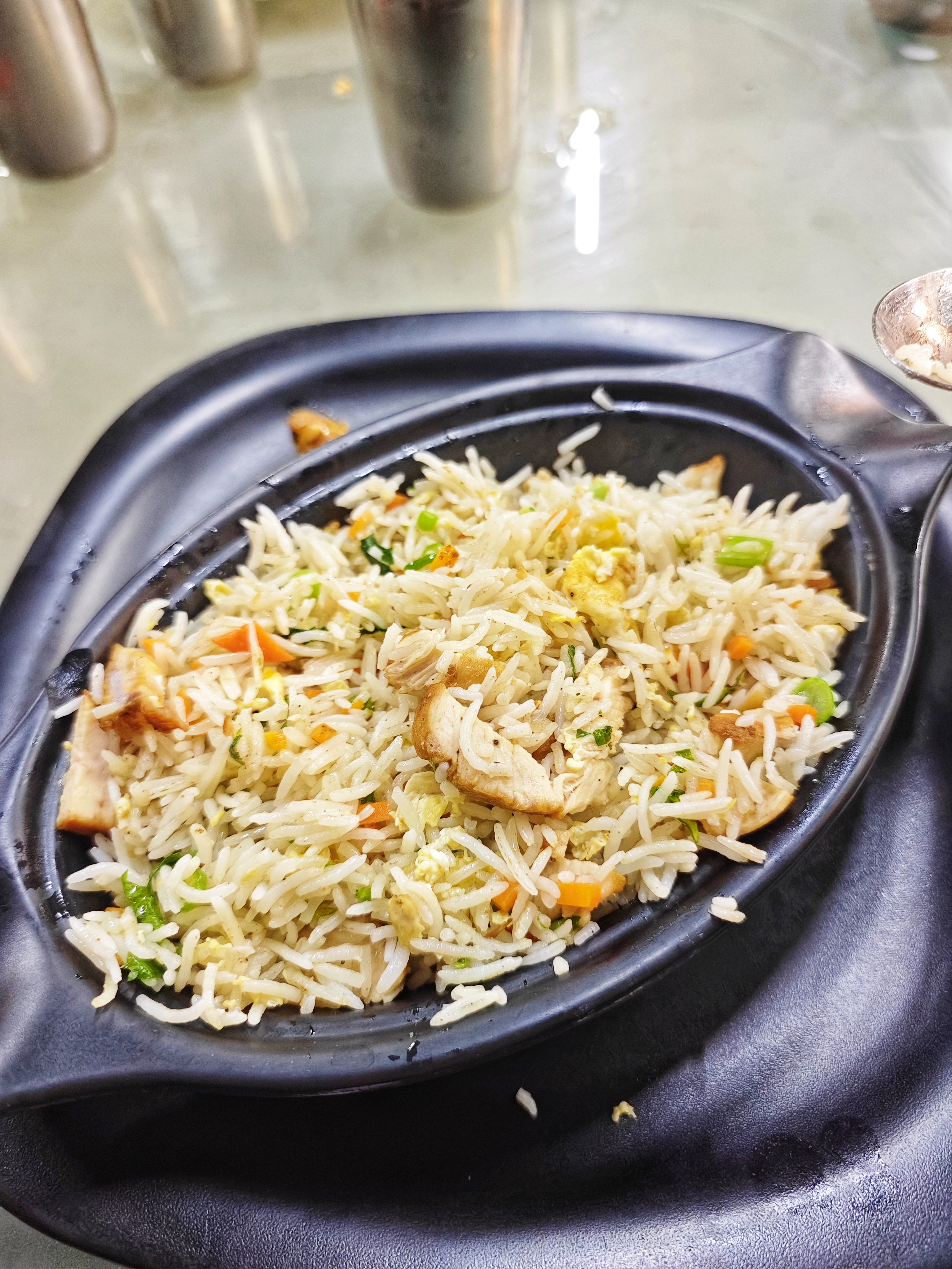 A black oval plate filled with chicken fried rice, featuring vibrant vegetables like carrots and peas, sits on a reflective table, evoking a warm, appetizing feel.