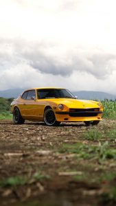 Low-angle three-quarter shot of a classic, bright yellow Datsun 260Z sports car , parked on a dirt and grass field in Costa Rica. The static car faces forward and slightly right under a dramatic, cloudy sky, with soft layers of distant mountains framing the natural backdrop.