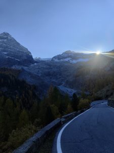 An asphalt mountain pass road curving to the right under a blue twilight sky. In the background, rocky, snow-dusted mountains rise above a valley filled with autumn-colored pine trees, with the bright sun bursting over a ridge on the right.
