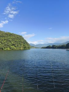 
Calm Fewa lake scene with fishing rods in foreground, surrounded by lush green hills under a clear blue sky