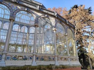 Detail of the Crystal Palace in Madrid. A facade of iron and glass with white arches and columns, decorated with colorful tiles featuring mythological motifs on the base and the frieze. Trees with autumn leaves can be seen in the background under a clear sky.