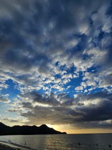 Dramatic gray clouds fill the sky above a dark shoreline and calm ocean water, with sunlight glowing behind a row of hills