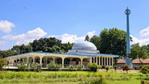 Rajshahi University Central Mosque surrounded by green lawns, trees, and a bright blue sky with white clouds on a sunny day.
