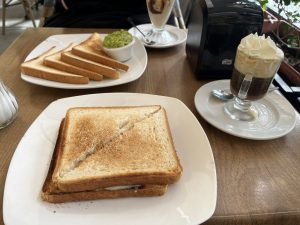 A wooden table in a café setting features a toasted sandwich, crispy toast with guacamole, and a dark drink topped with whipped cream. A toaster and a dessert in the background complete the cozy atmosphere.