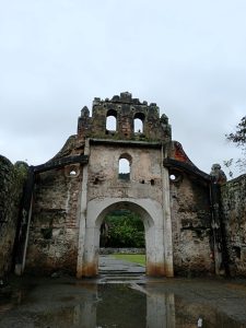 View from inside the ruins of the Ujarrás church in Costa Rica, looking out through the crumbling stone facade with its arched doorway and bell-gable, framed by tropical vegetation.
