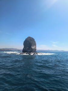 
Large rock formation rising from the blue sea with green hills behind.