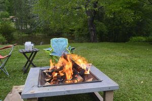 Outdoor fire pit with burning logs surrounded by camping chairs on a grassy lawn.