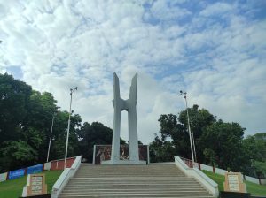 A wide low-angle shot of the modern concrete Shaheed Minar monument at Rajshahi University of Engineering and Technology (RUET), featuring a wide staircase leading up to the structure under a bright blue sky filled with textured white clouds.