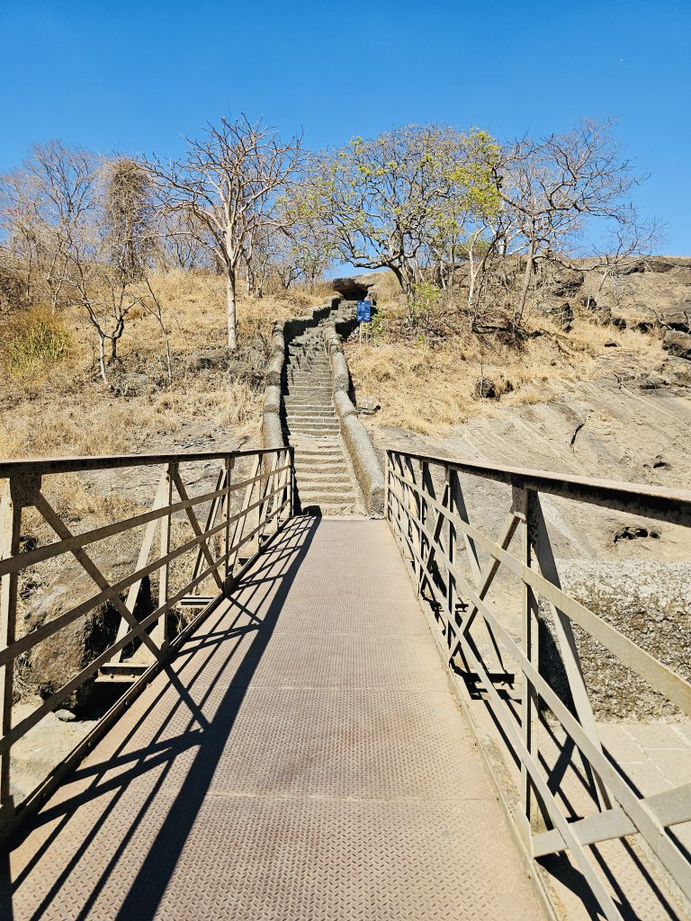 A metal bridge leads to a long stone staircase surrounded by dry forest, inviting visitors to explore the higher caves. The clear blue sky adds depth to the peaceful landscape. Captured at Kanheri Caves, Borivali East, Mumbai.