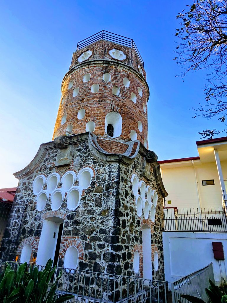 Tall round brick tower with numerous white-rimmed niches, built atop a stone base with additional oval niches, set against a clear blue sky with a tree branch in the foreground.
