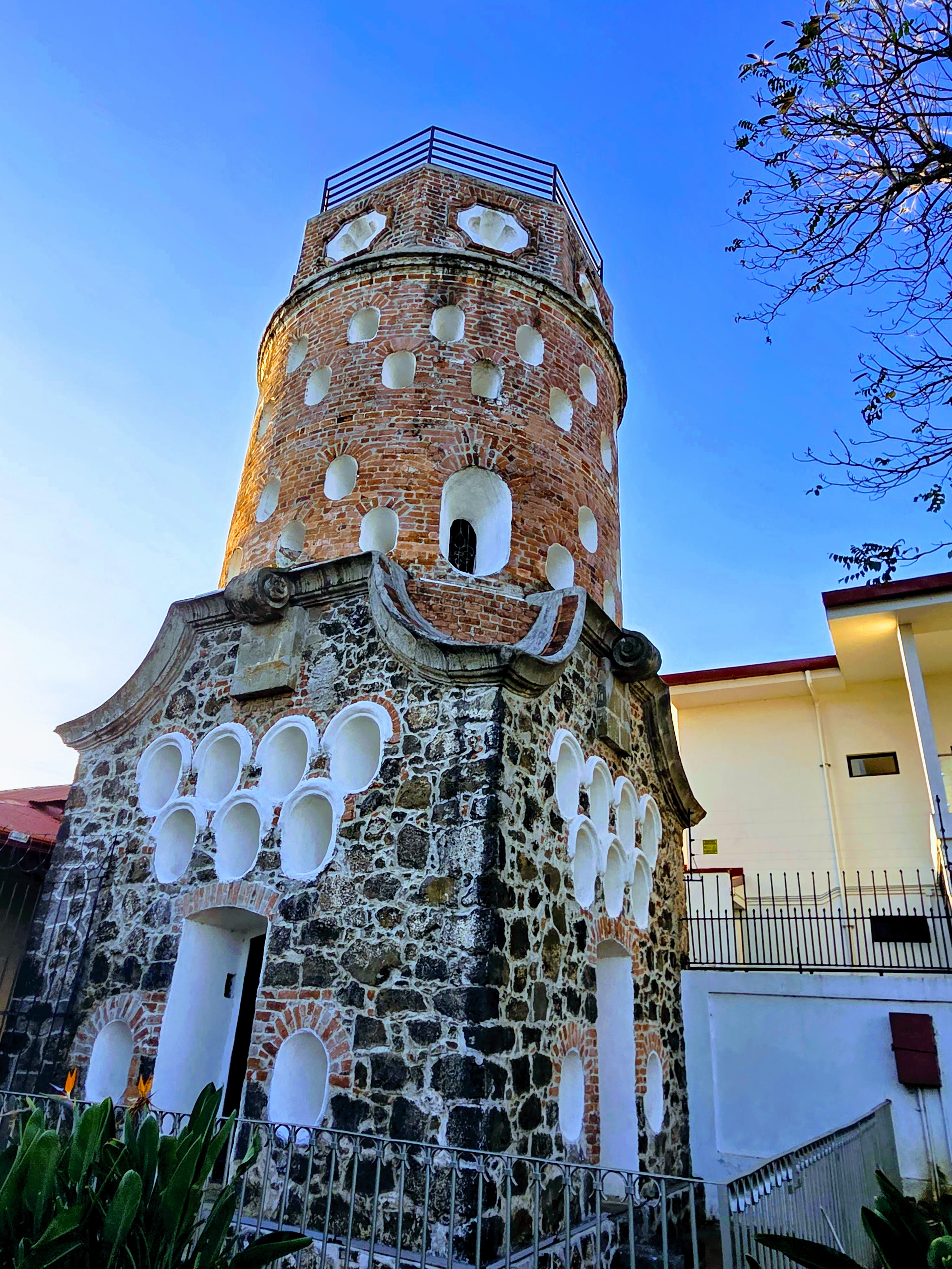 Tall round brick tower with numerous white-rimmed niches, built atop a stone base with additional oval niches, set against a clear blue sky with a tree branch in the foreground.