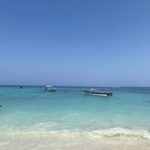 Clear turquoise water at a sandy beach with small boats anchored under a bright blue sky.