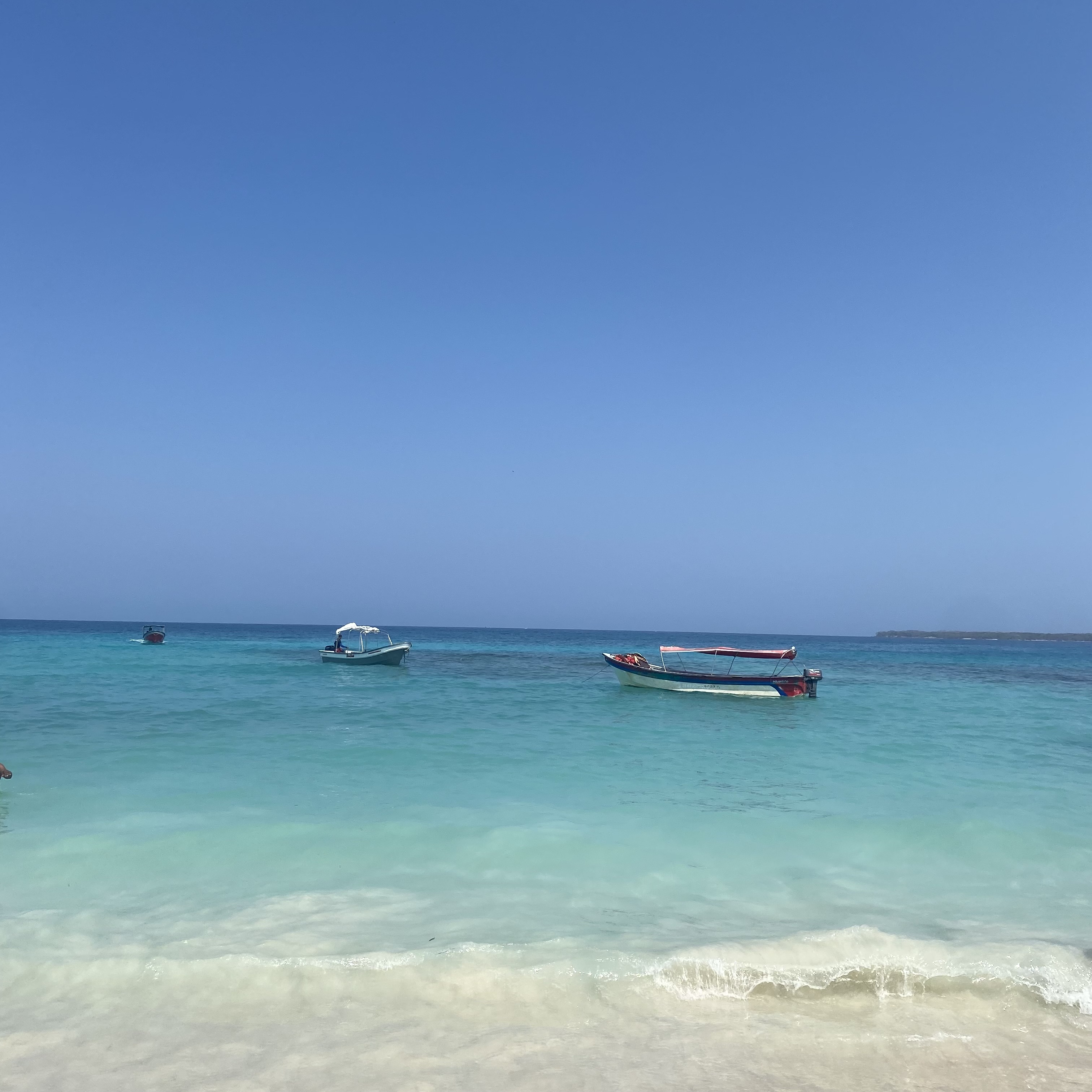 Clear turquoise water at a sandy beach with small boats anchored under a bright blue sky.