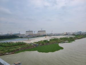 An elevated view shows a large industrial plant under construction with two cooling towers next to a wide river under a cloudy sky.
