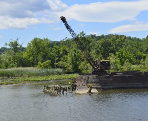 A large old crane sits on a barge in a calm river, surrounded by green trees and vegetation under a partly cloudy blue sky