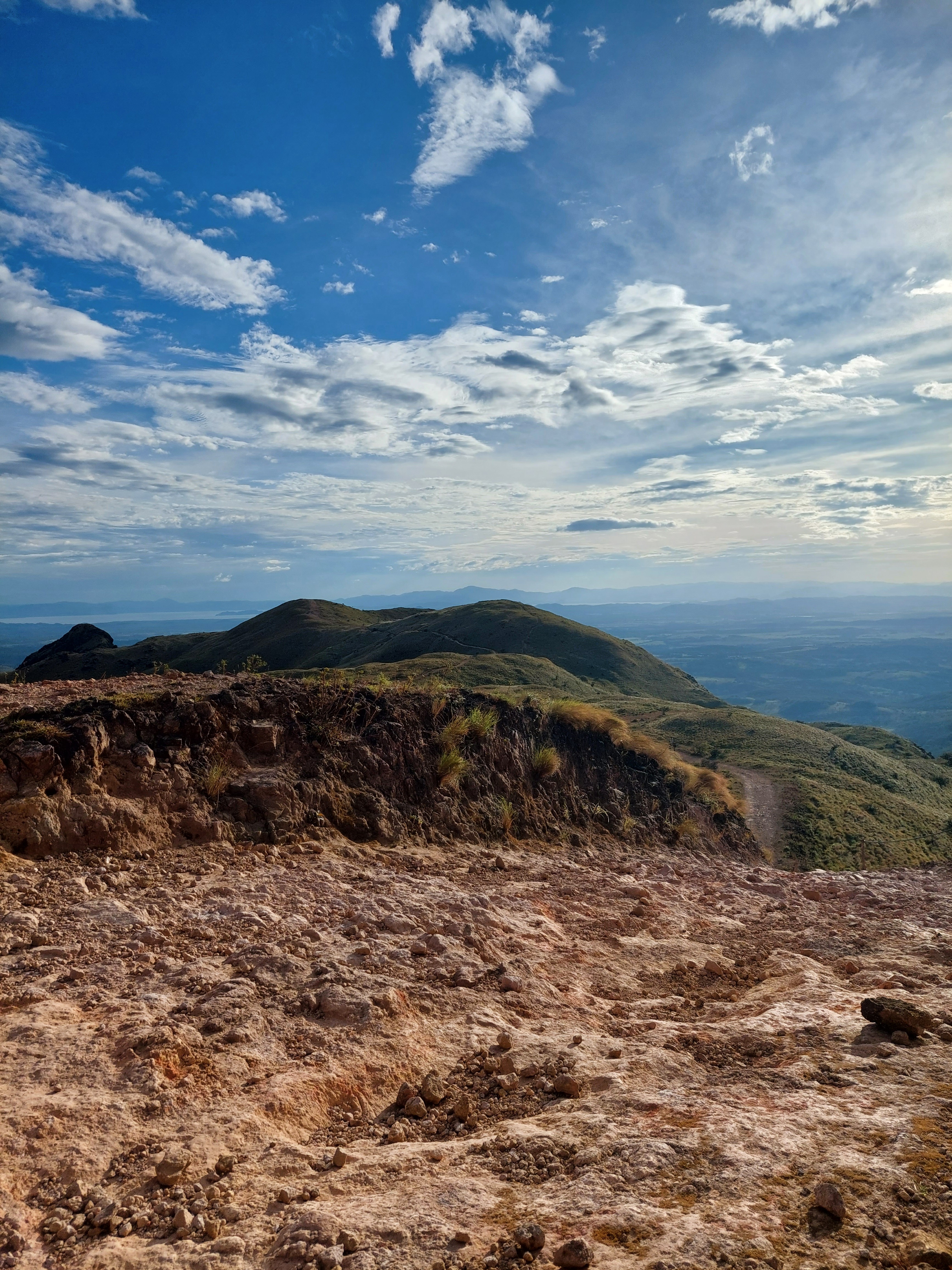 Rocky terrain overlooks rolling green hills beneath a wide blue sky filled with large, fluffy clouds. The open mountain landscape creates a sense of depth and highlights the natural beauty of the surrounding countryside