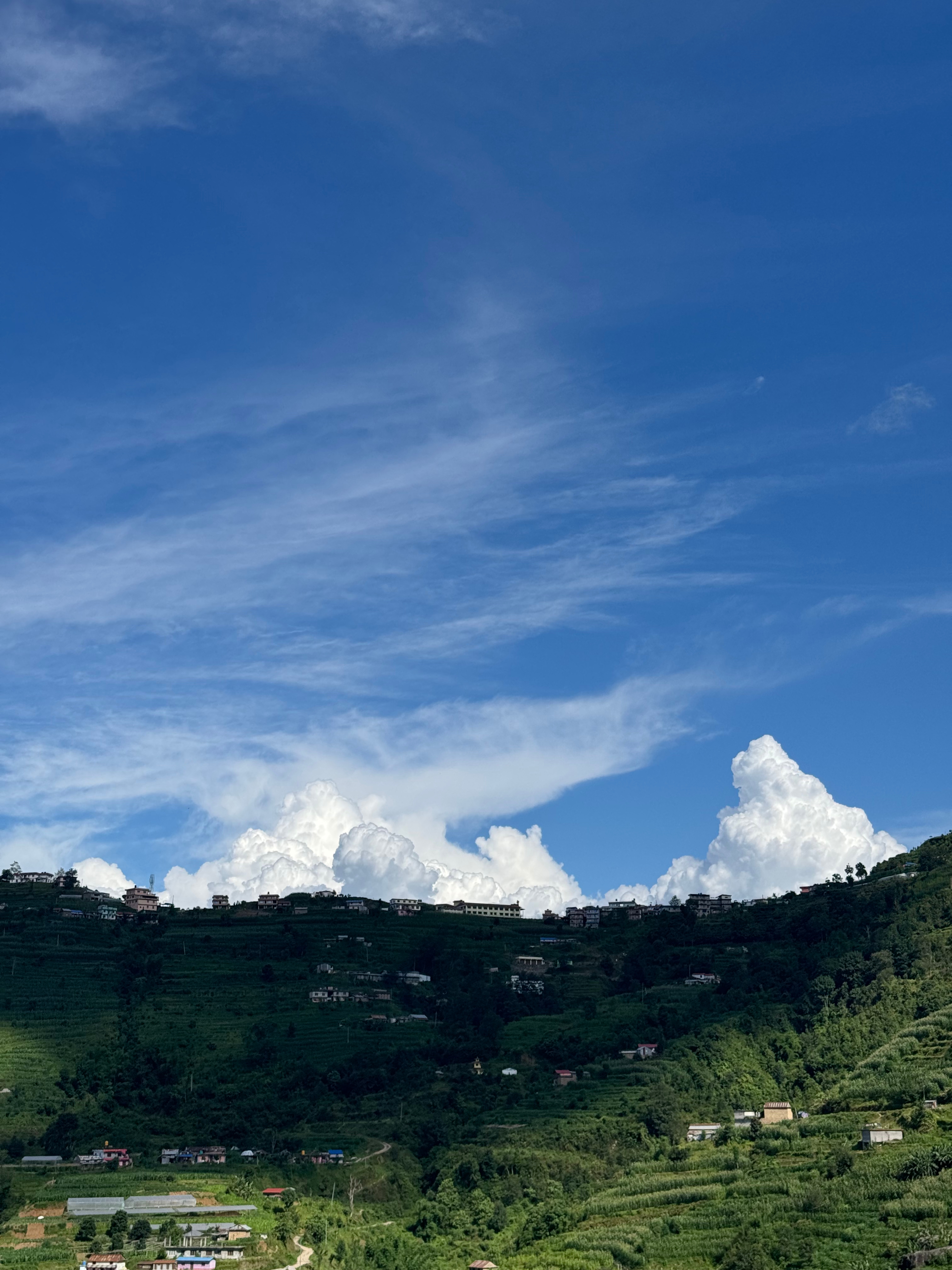 A lush green hillside with scattered houses under a vibrant blue sky filled with fluffy white clouds