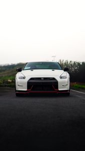 A low-angle front view of a white Nissan GT-R R35 Nismo sports car positioned on the asphalt of a rain-slicked road in the mountains of Costa Rica. Green, tree-covered hills form the background beneath a bright sky with diffused light.