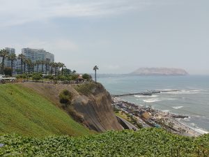 A coastal landscape with a green cliff, beachfront promenade, pier, sandy beach, ocean waves, and buildings with palm trees under a clear blue sky.