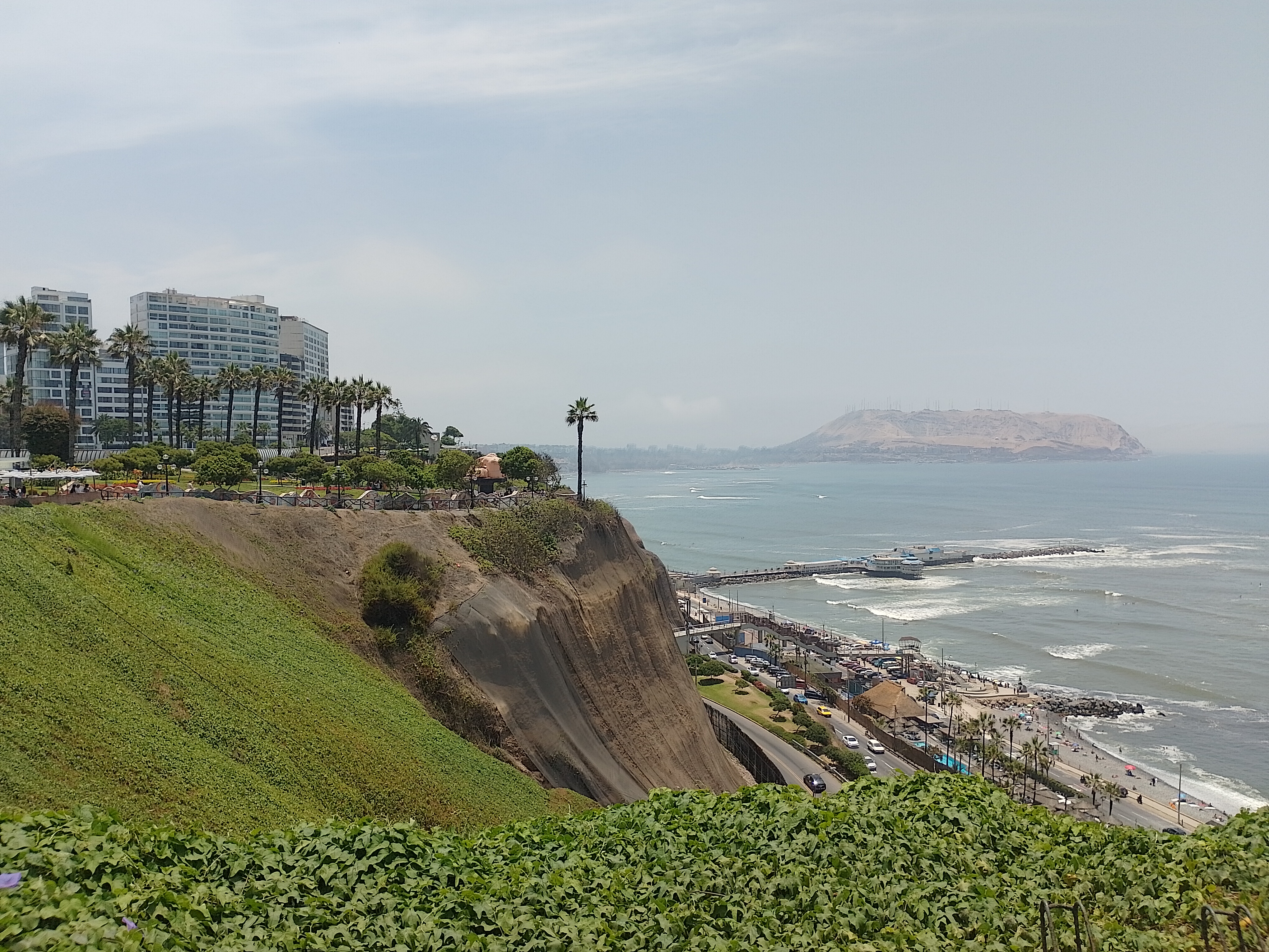 A coastal landscape with a green cliff, beachfront promenade, pier, sandy beach, ocean waves, and buildings with palm trees under a clear blue sky.