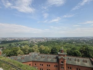 Aerial view from a red-brick fortress or historic building with a corner tower, overlooking a green forested park and a sprawling city skyline under a partly cloudy blue sky.
