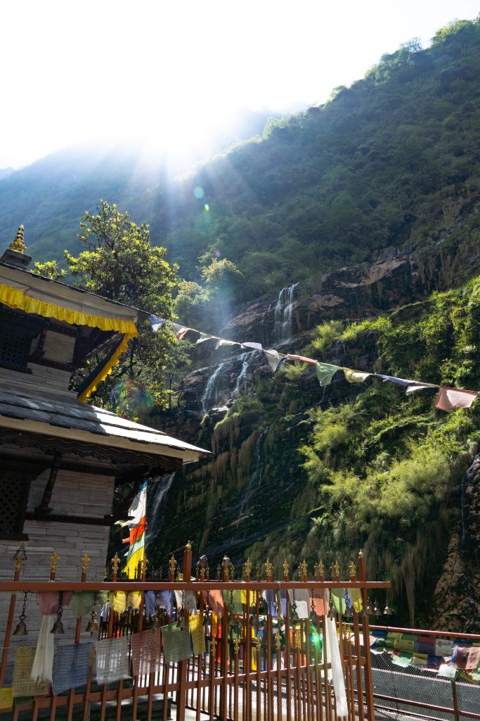 Buddhist temple with pagoda roof and prayer flags on a metal fence, waterfalls streaming down a green mountainside in the background