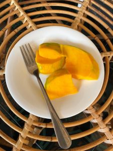 Slices of ripe yellow mango on a white plate with a fork, set on a woven rattan surface. 
