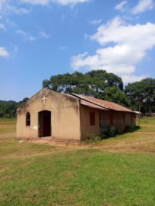 A small rural church with a weathered corrugated iron roof, surrounded by grass and large trees under a blue sky with scattered clouds.
