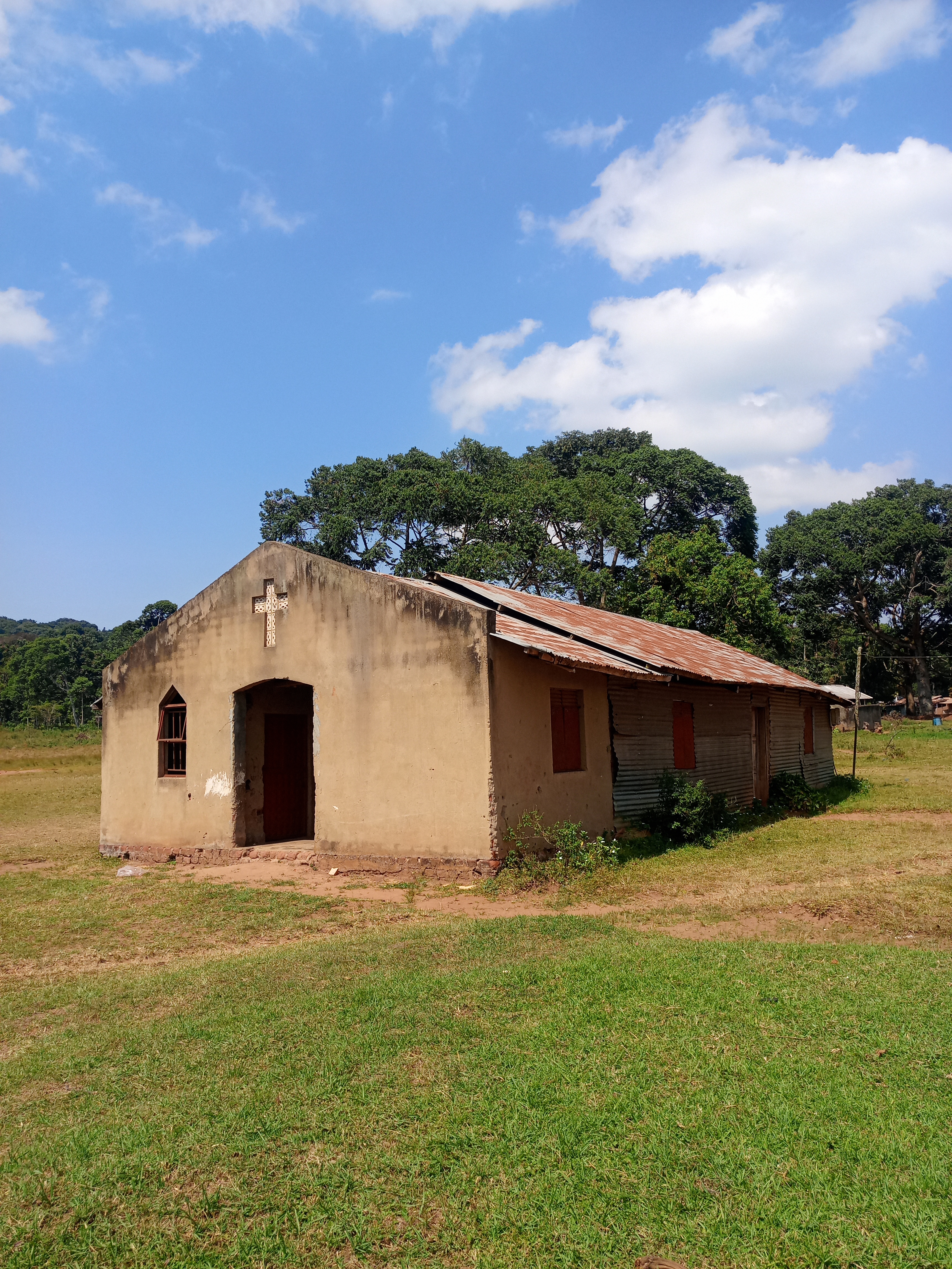 A small rural church with a weathered corrugated iron roof, surrounded by grass and large trees under a blue sky with scattered clouds.