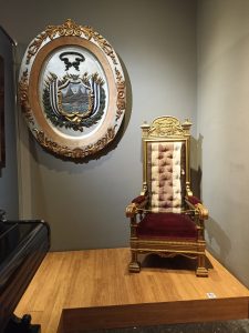 Ceremonial presidential chair in red velvet and gold detailing, displayed alongside a historic Costa Rica coat of arms at the National Museum of Costa Rica in San José.